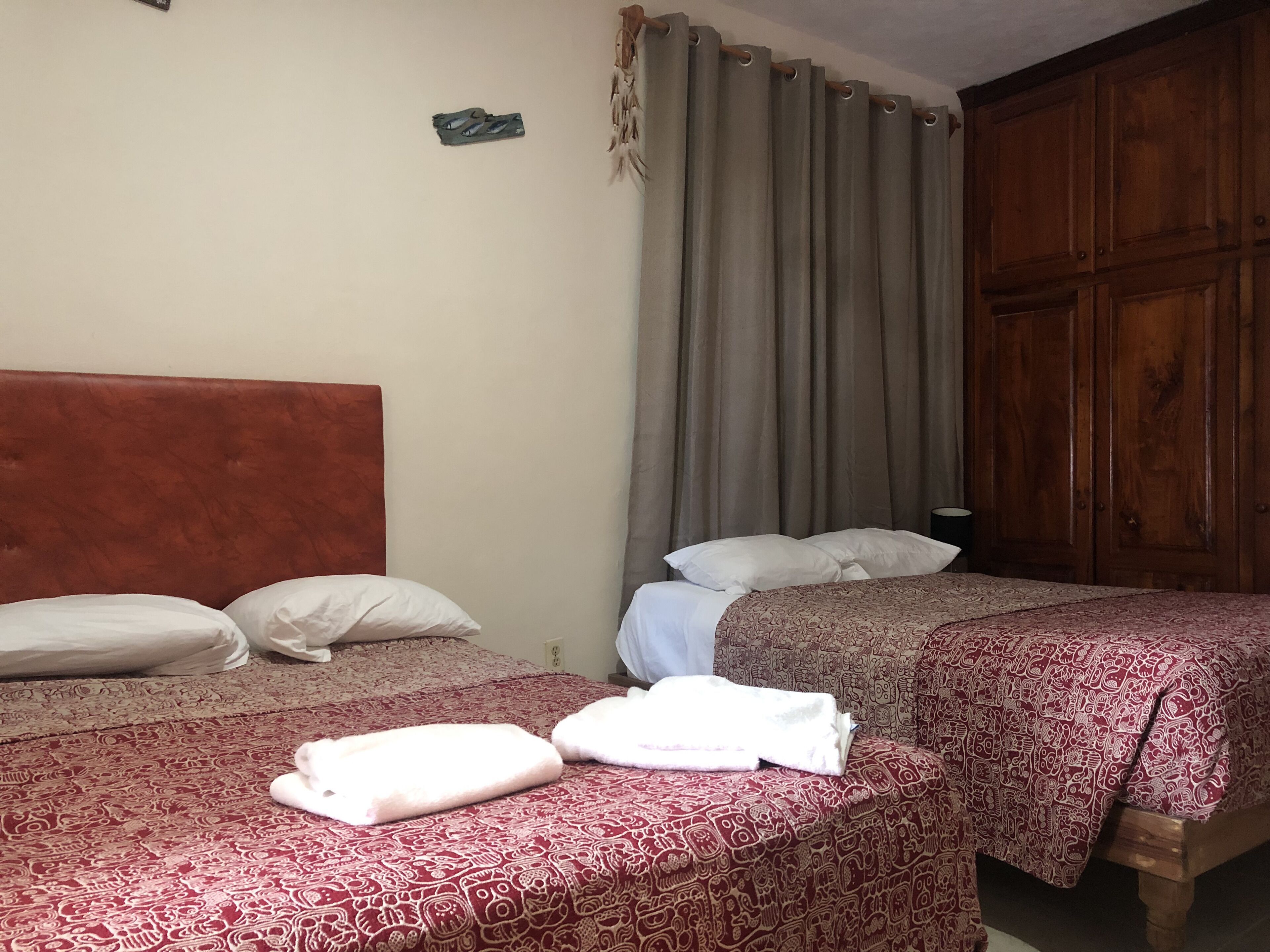 Une chambre avec deux lits, chacun agrémenté d’oreillers blancs et d’une couverture à motifs rouges. On y trouve une armoire en bois et un rideau à la fenêtre.