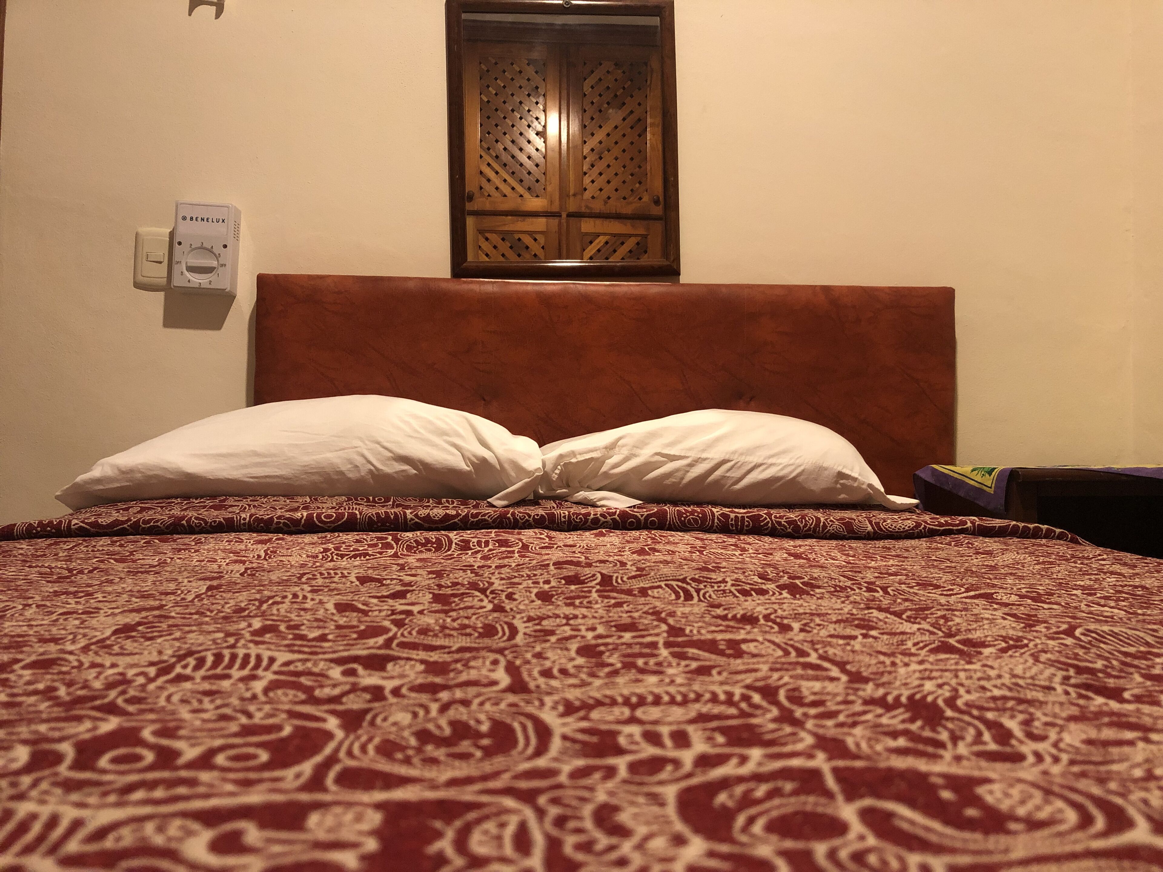 A bed with a patterned bedspread, two pillows, a wooden headboard, a wall-mounted thermostat, and a window with a wooden lattice design.