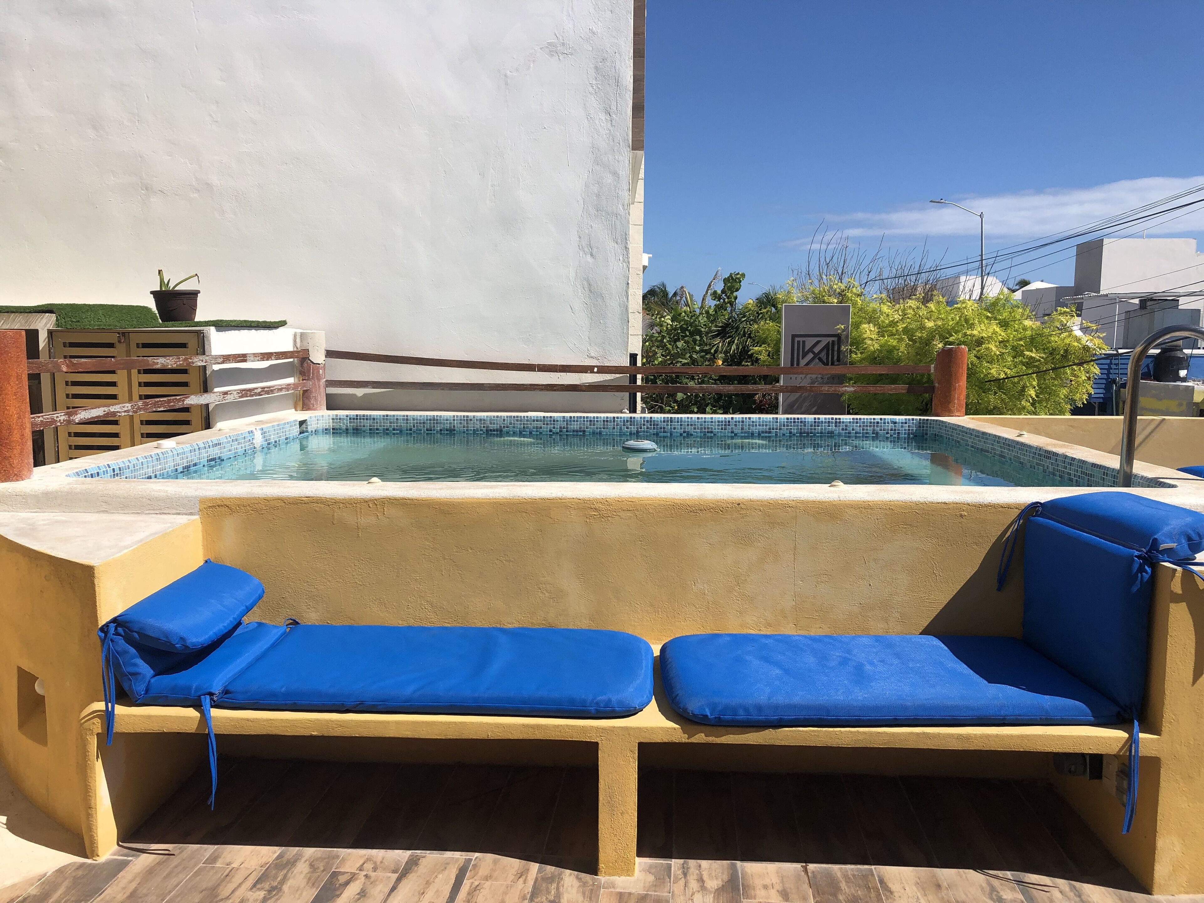 Una piscina en la azotea con sillas acolchadas azules, un borde de azulejos y vistas a edificios y áreas verdes.