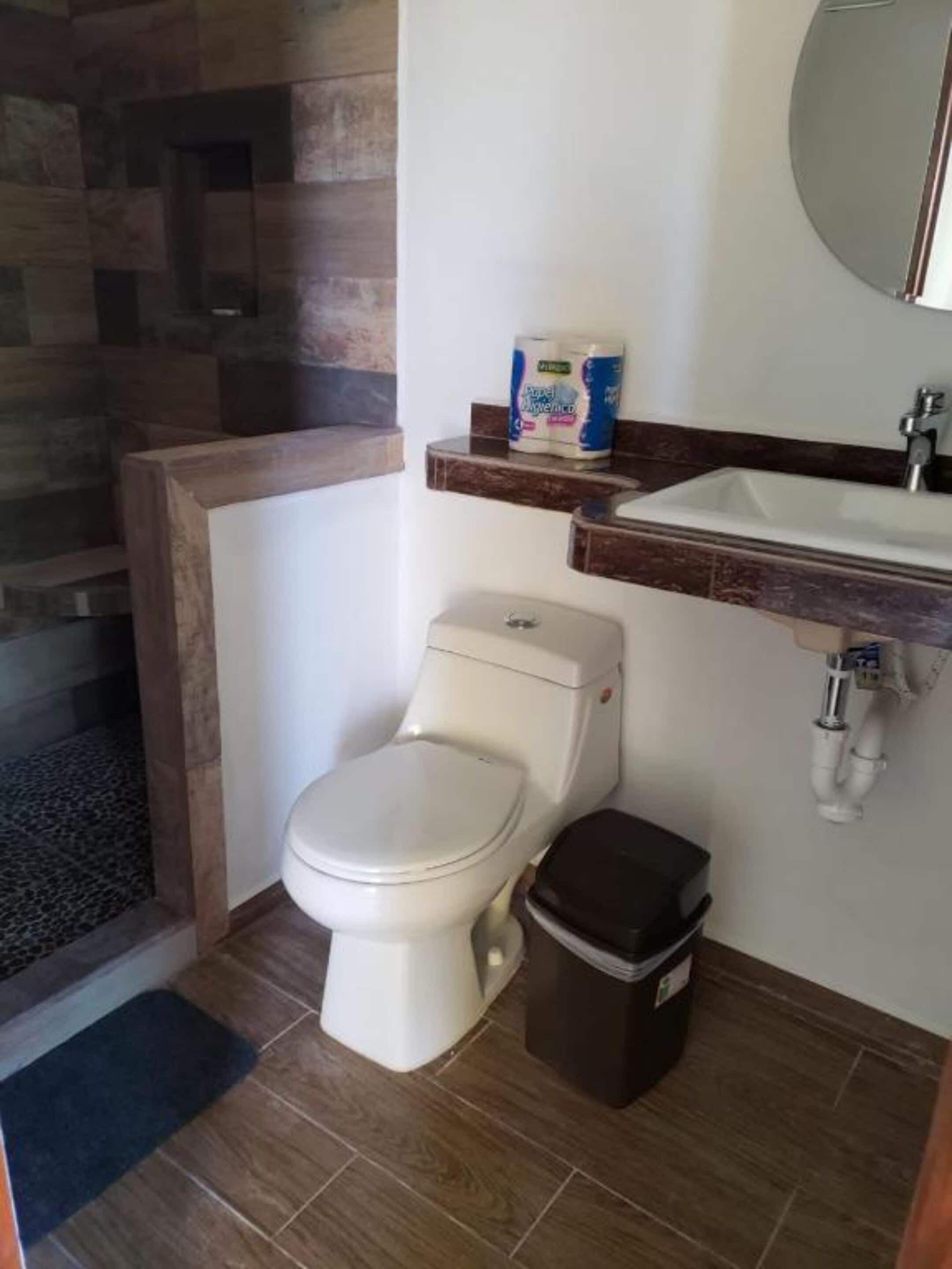 Uma casa de banho com sanita, um contentor de lixo, lavatório e dispensador de papel higiénico.