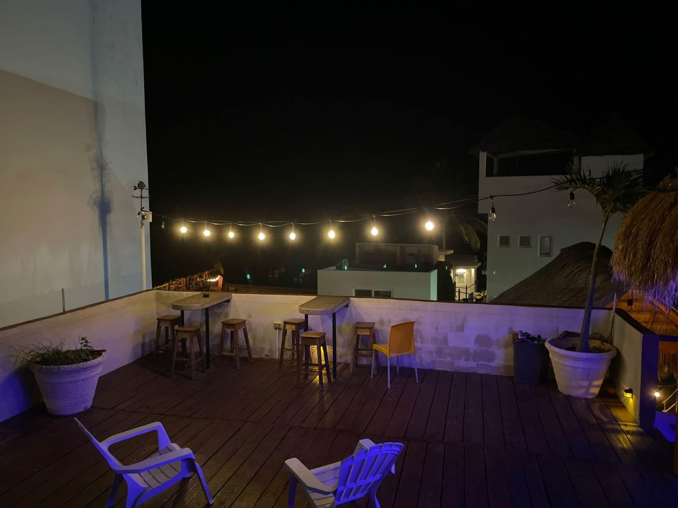 A rooftop terrace with wooden flooring, several chairs, a small table, string lights, and potted plants.