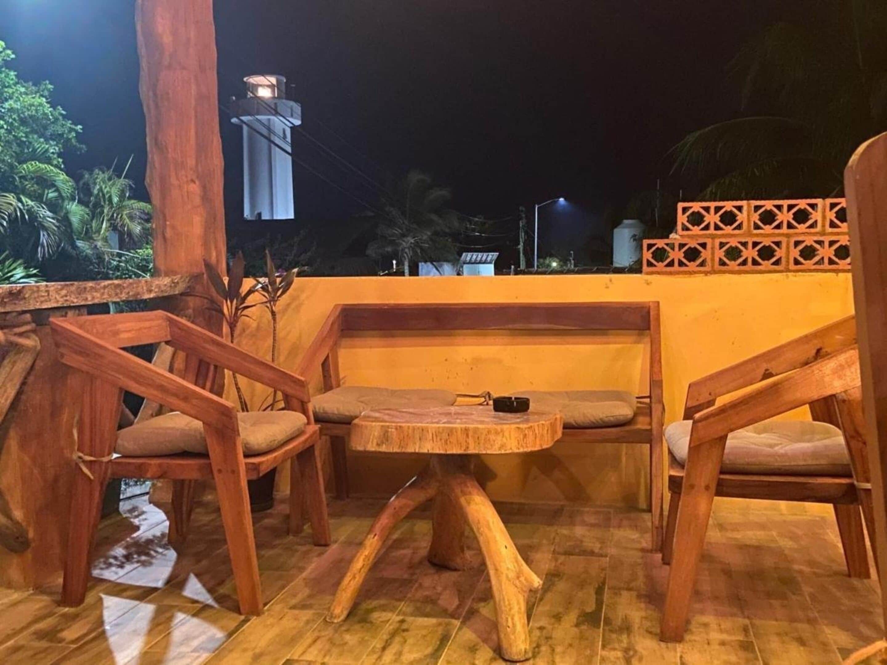 A wooden outdoor seating area with a table and chairs, a small table lamp, and a decorative wall.