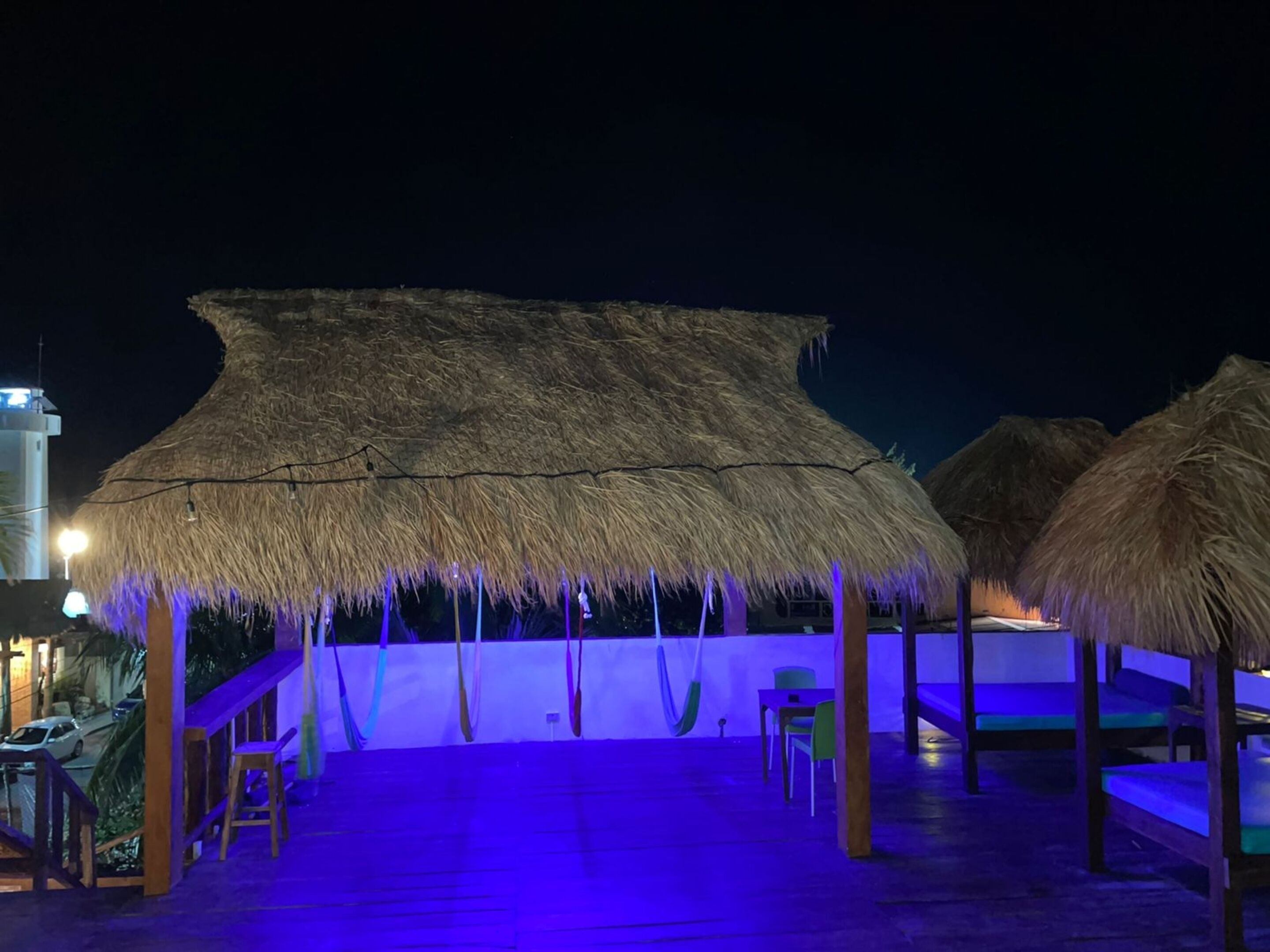 A thatched-roof outdoor area with wooden benches and tables, a staircase, and a view of a building with a tower.