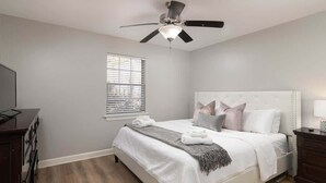 Ein Schlafzimmer mit einem Bett, einem Deckenventilator, einem Fenster mit Jalousien, einem Schminktisch und einem Nachttisch mit Lampe.
