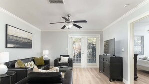 Ein modernes Wohnzimmer mit Deckenventilator, schwarzen Ledersofas, einem Glastisch, einem Fernseher und einer Schiebetür zu einem Außenbereich.