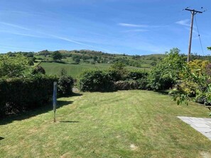 A grassy backyard with a stone pathway leading to a gate, surrounded by lush greenery and hills in the background.