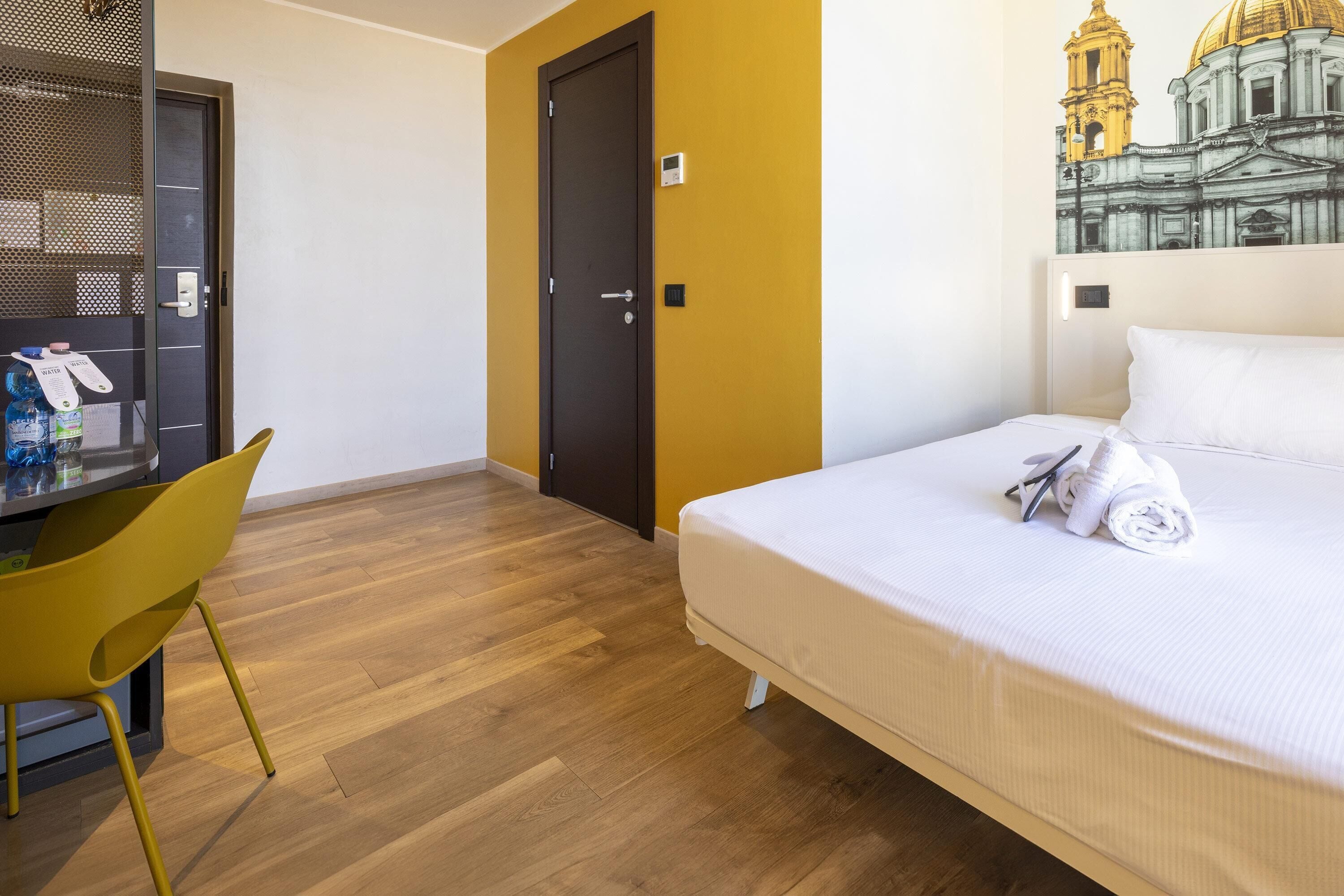 Ein Hotelzimmer mit einem Bett, einem Schreibtisch mit Stuhl, einer Tür und einer Wand mit Wandgemälde.