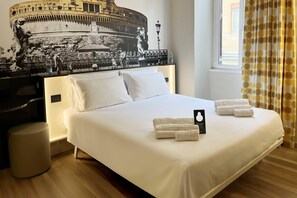 A hotel room with a large bed, white bedding, a headboard with a mural, a bedside table, and a window with curtains.