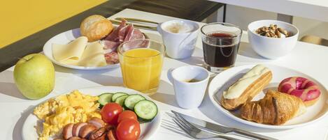 A breakfast table with plates of eggs, sausages, tomatoes, cucumbers, a croissant, and a glass of orange juice.
