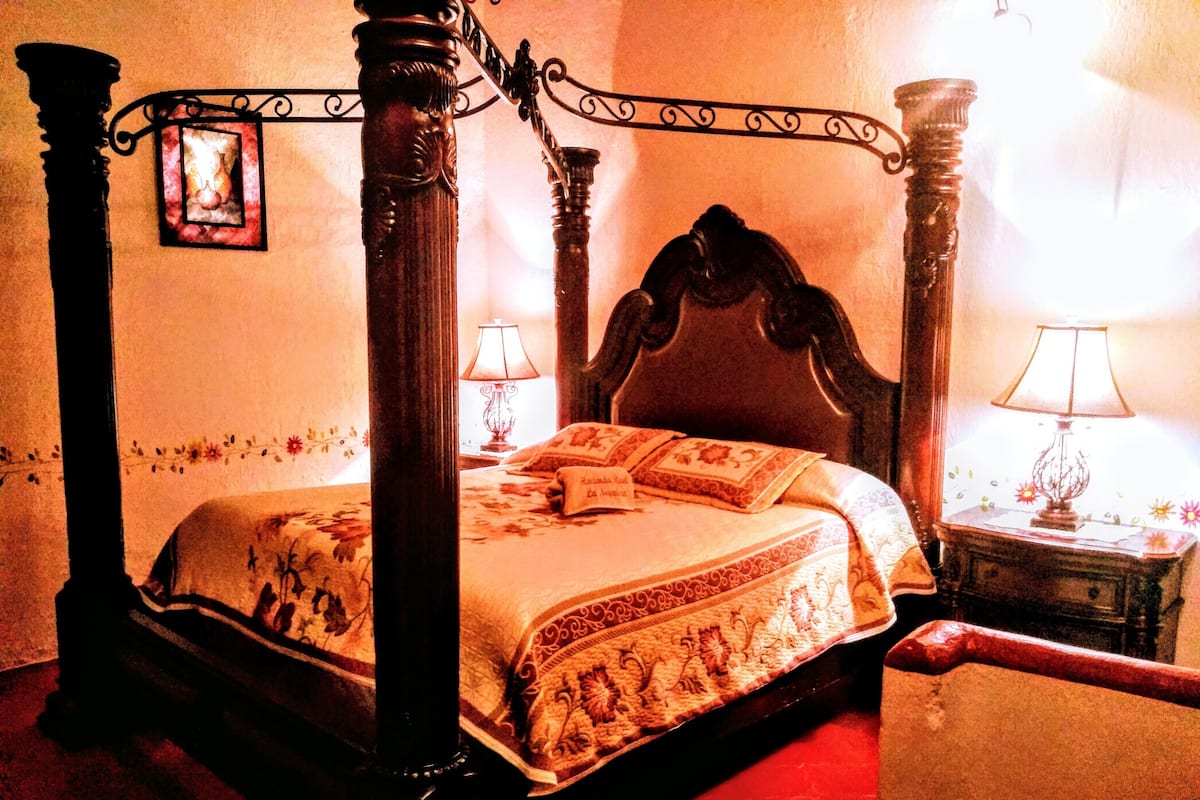 Ein Himmelbett mit aufwendigen Holzsäulen und einem geschnitzten Kopfteil, begleitet von zwei Nachttischen mit Lampen.