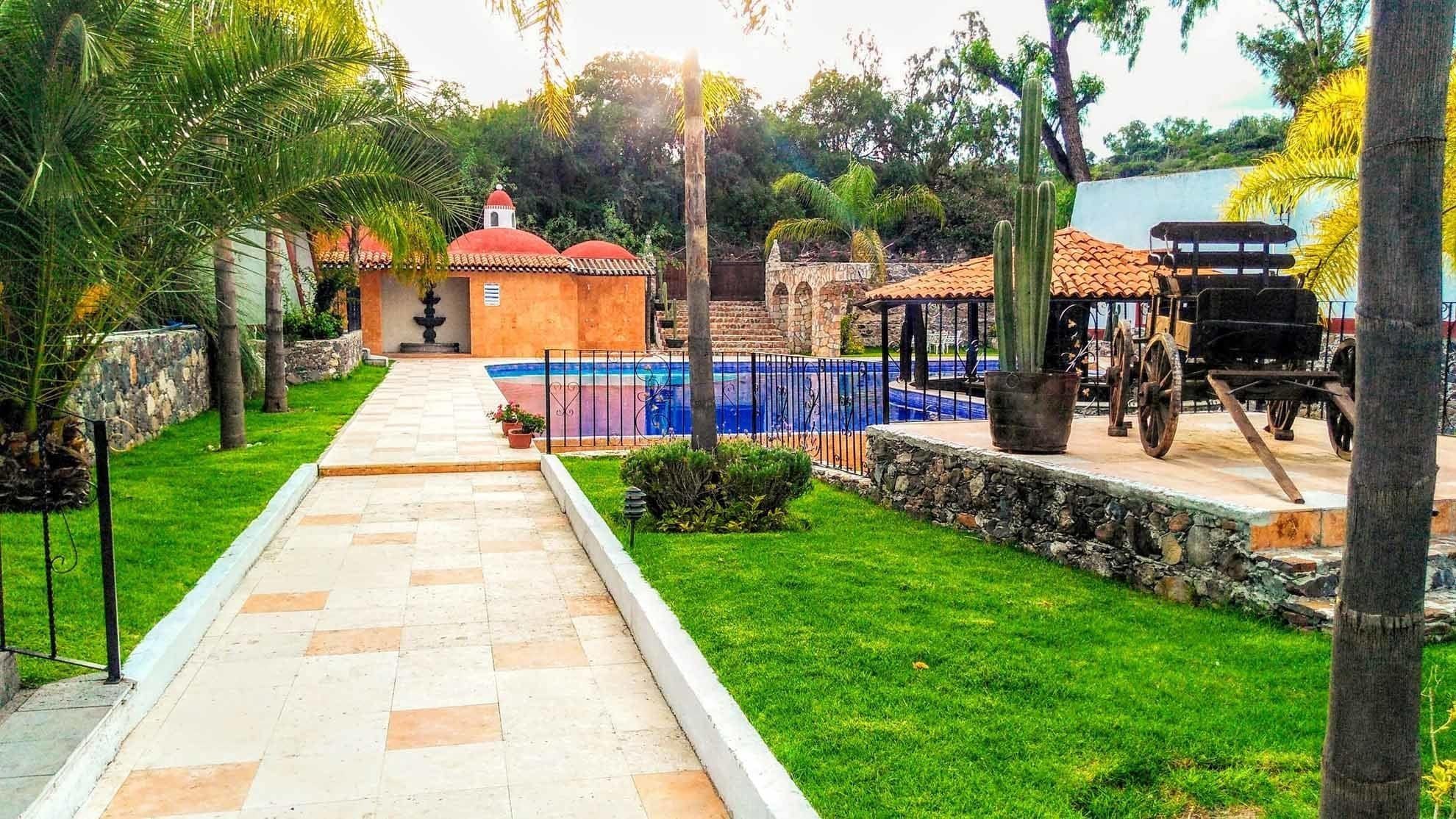 Un sendero conduce a un área de piscina con una fuente, rodeada de vegetación y un muro de piedra.