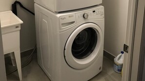A compact laundry room with two stacked front-loading washing machines, a small white table, and a bottle of detergent on the floor. - Premium Elite suites (Oshawa)