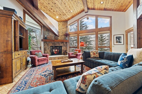 A spacious living room with a fireplace, large windows, and a high wooden ceiling.