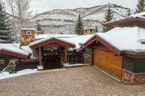 A snow-covered lodge with a stone chimney, a wooden garage door, and a paved driveway. - Cascade Village Private Mountain Home (Vail)