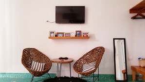 Une pièce comprenant deux fauteuils en osier, une petite table ronde, un téléviseur fixé au mur, une étagère avec des photos encadrées et un miroir.
