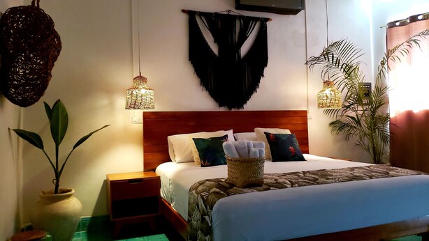 Une chambre à coucher bien aménagée, avec une tête de lit en bois, un grand lit agrémenté de plusieurs oreillers, une table de chevet sur laquelle repose un panier en osier, et deux luminaires suspendus.