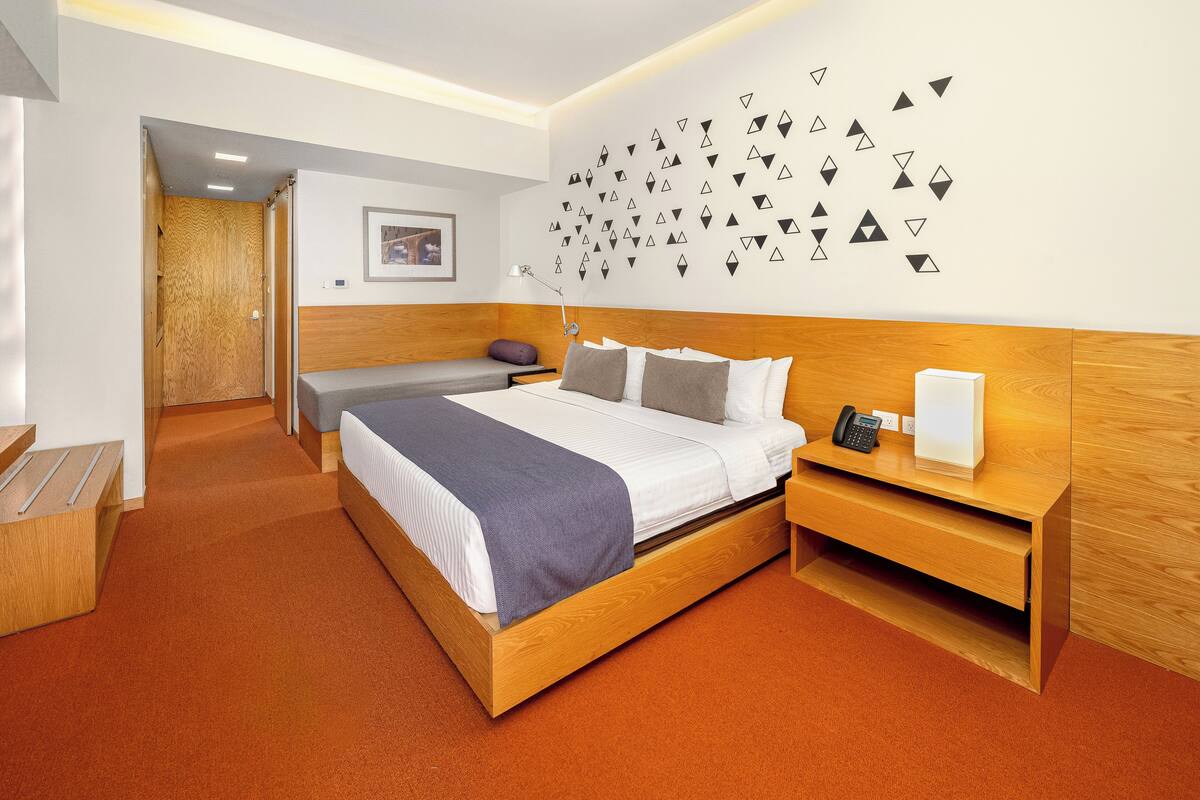 Ein modernes Hotelzimmer mit einem Holzbett, einem Nachttisch mit Telefon und Lampe, einer kleinen Sitzecke und einer Wand mit geometrischen Wandbildern.
