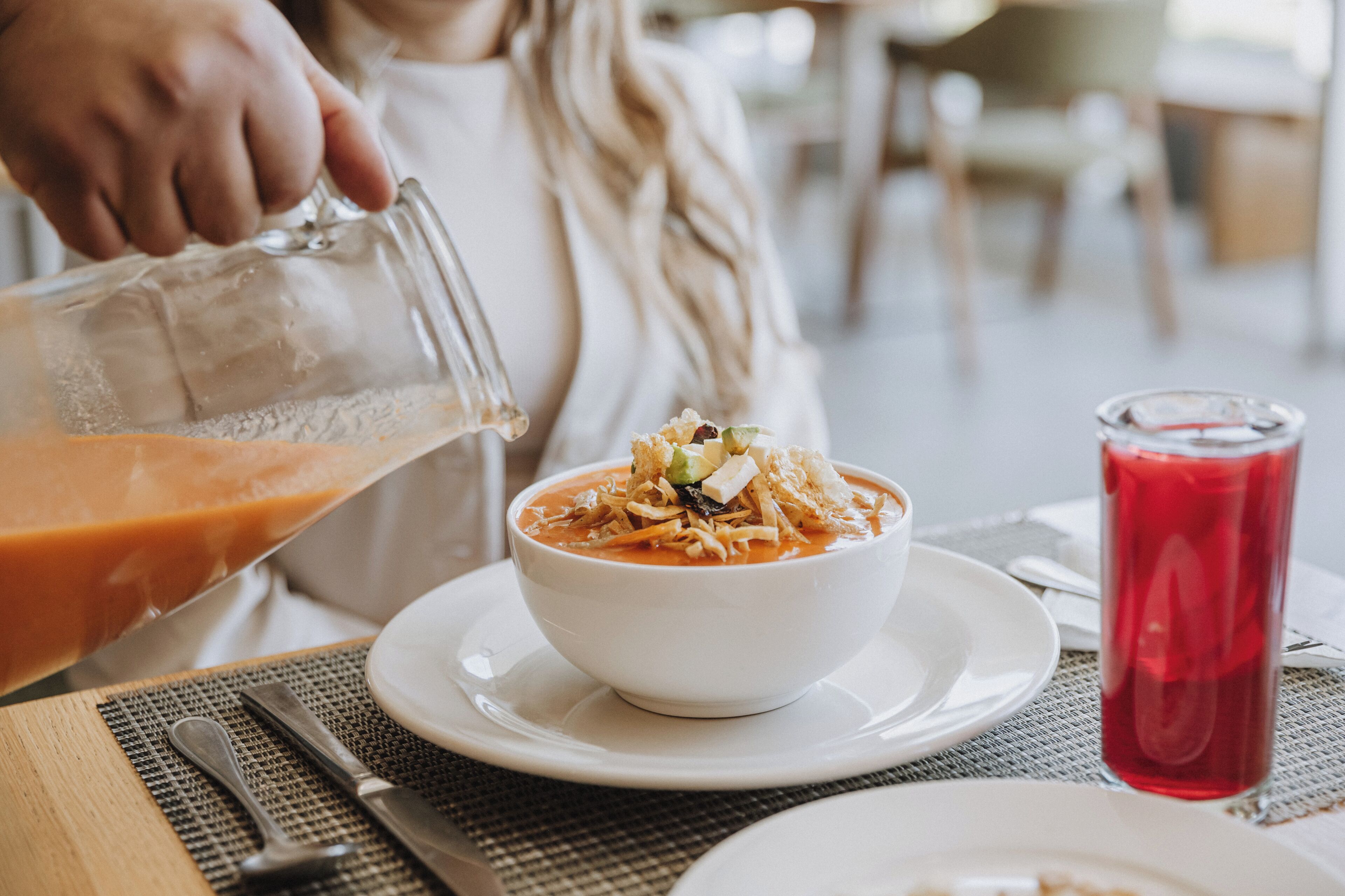 A dining table with a bowl of soup, a glass of red drink, and a person pouring a clear liquid into a glass.