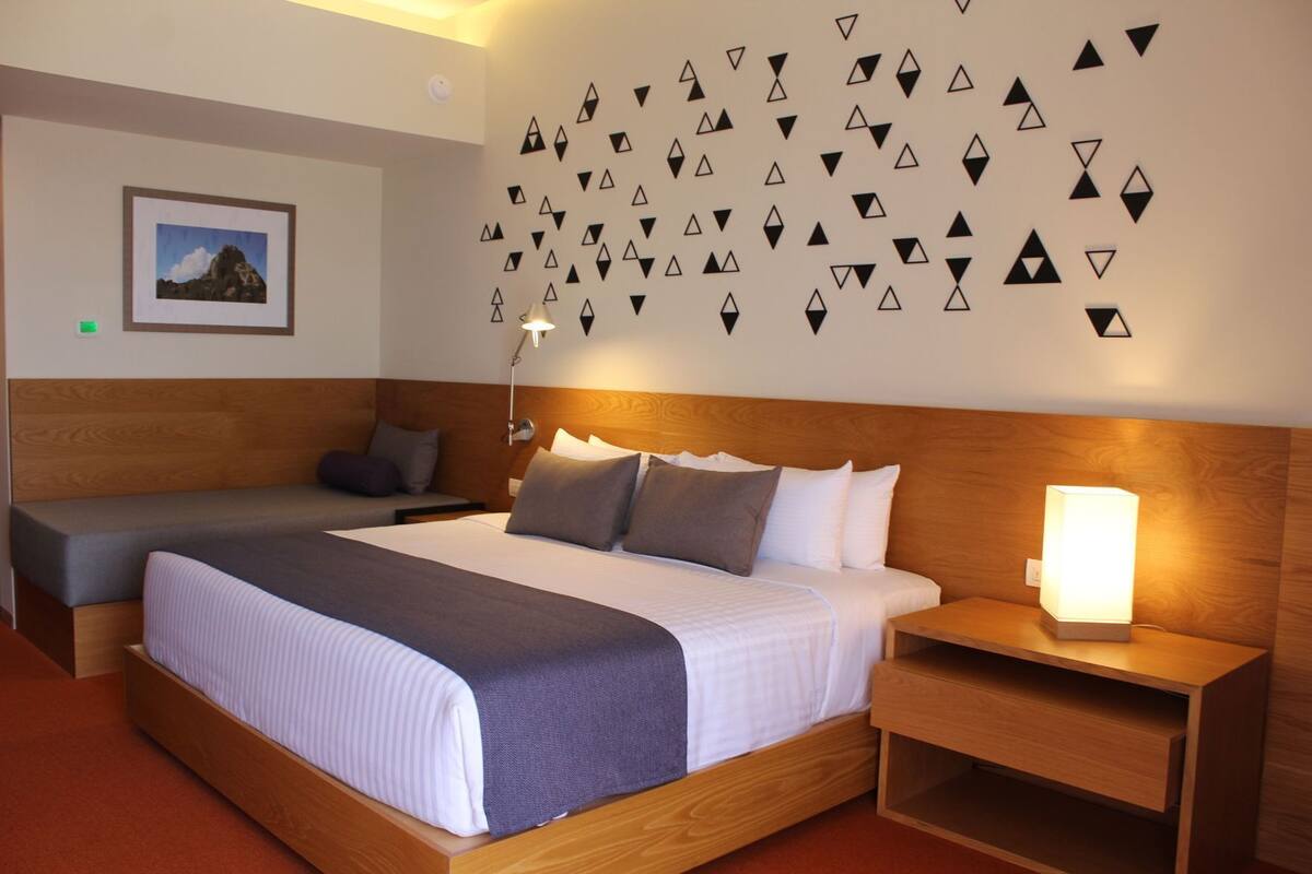 Ein Hotelzimmer mit einem großen Bett, einem kleineren Bett, einem Nachttisch mit Lampe, einem Wandbild und einer geometrischen Wanddekoration.