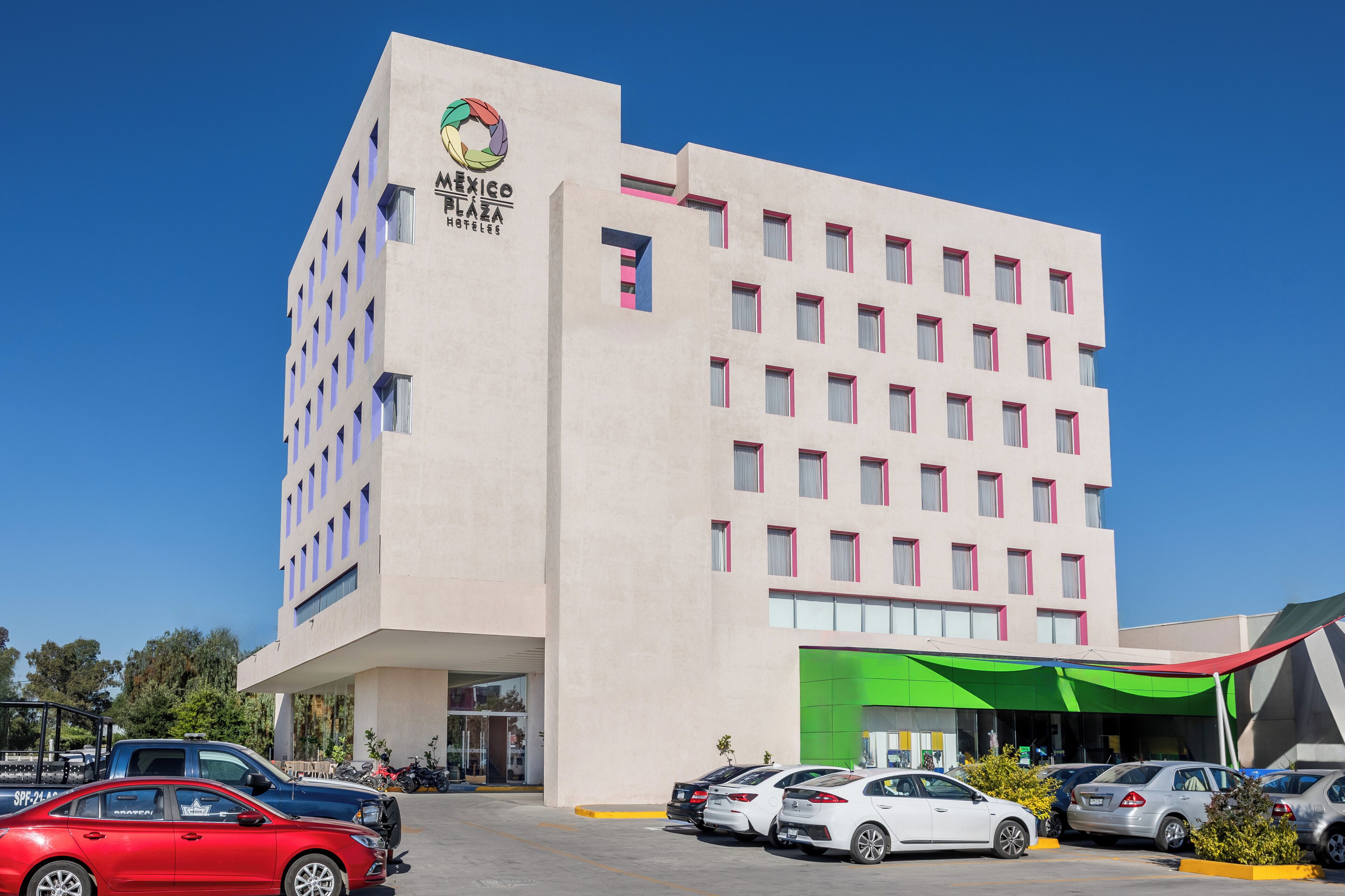 A multi-story hotel with a modern facade, a prominent logo, and a parking area with several cars.