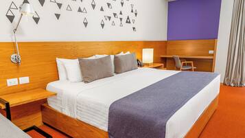 A hotel room with a large bed, a desk, a chair, a lamp, and a wall with geometric patterns.