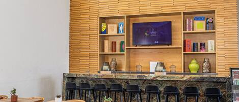 Zona de bar moderna con pared de madera, un televisor de pantalla plana y varios objetos decorativos. Hay varias opciones de asiento, incluyendo un sofá y mesas.