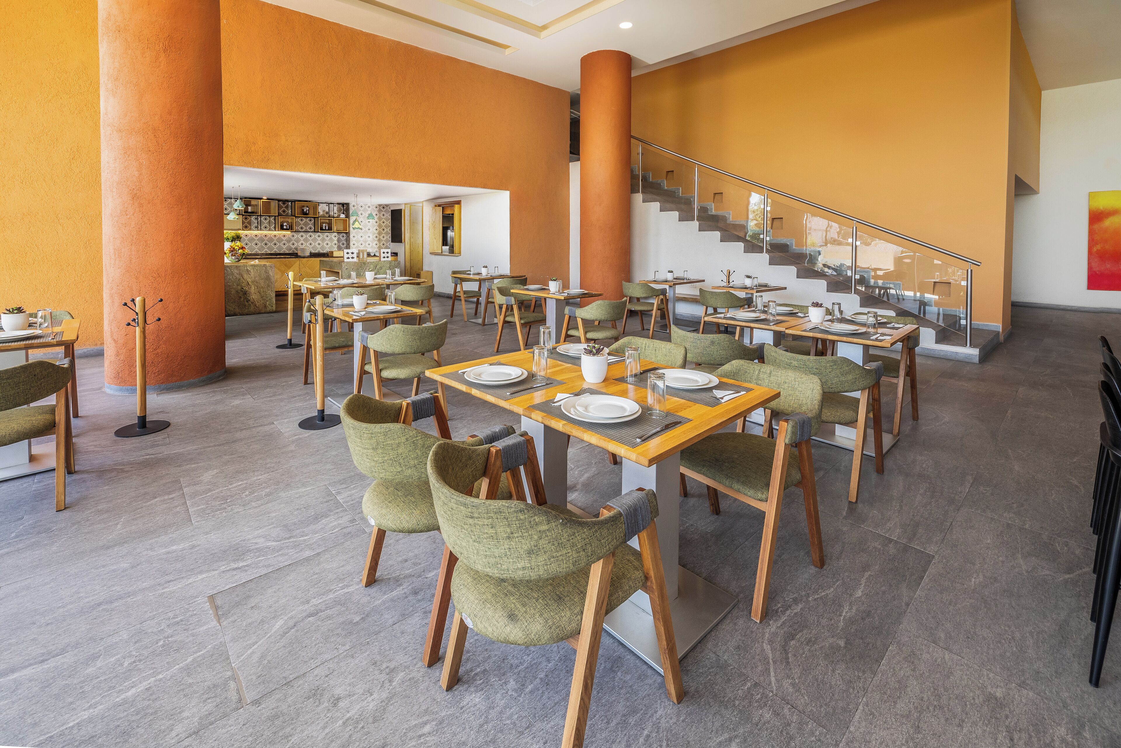 A spacious dining area with multiple tables and chairs, a staircase, and a kitchen in the background.