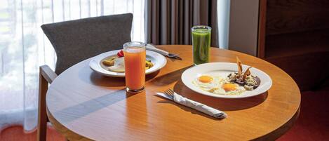 Habitación de hotel con una mesa redonda de madera dispuesta para el desayuno, incluyendo dos platos de comida y un vaso de jugo de naranja.