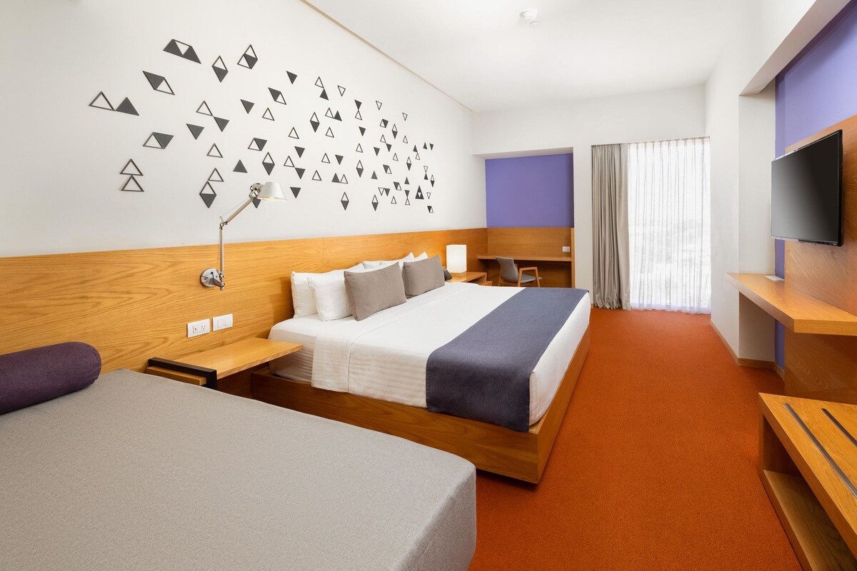 A hotel room with a bed, a desk, a TV, and a wall with geometric patterns.