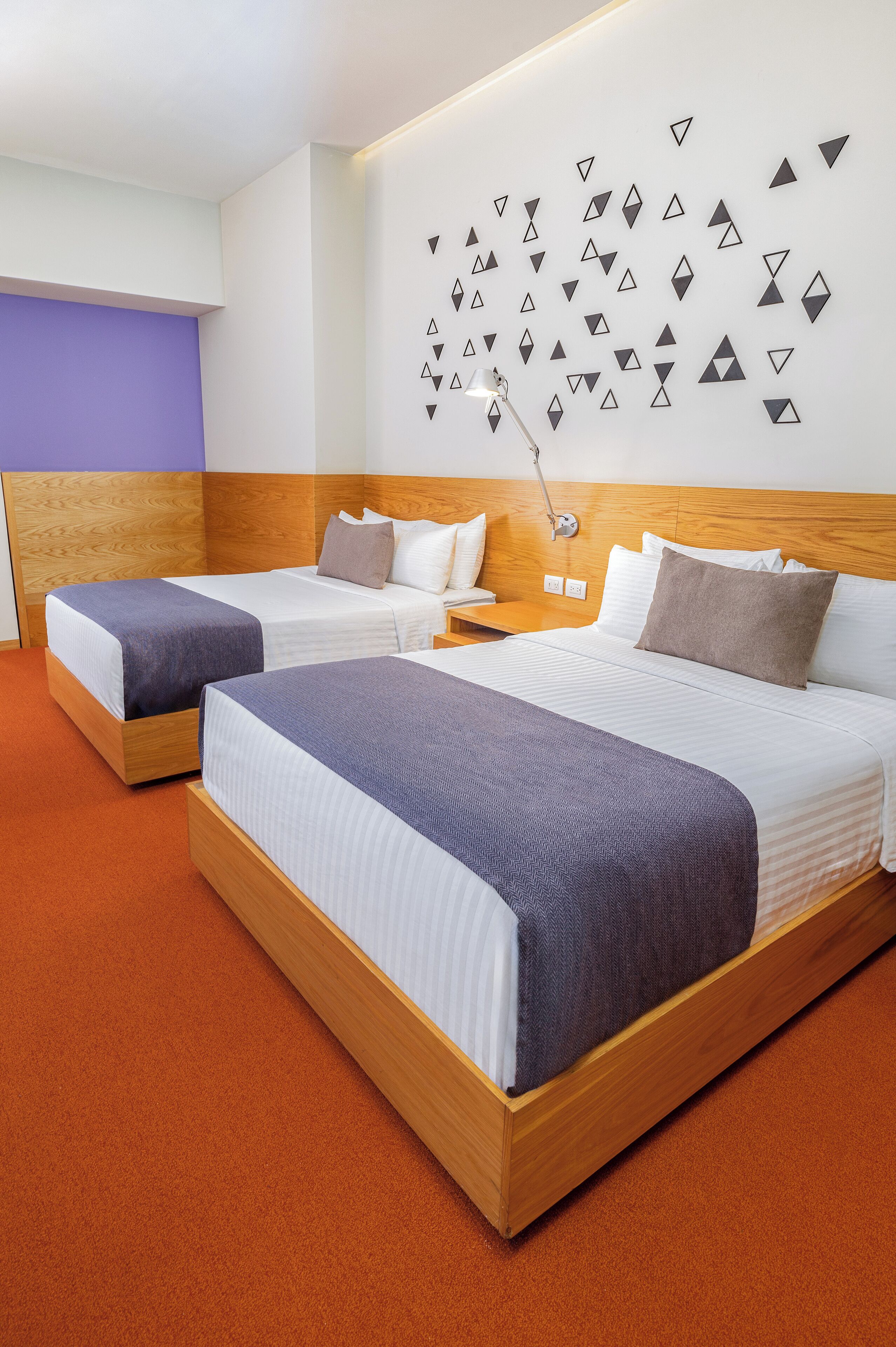 A hotel room with two beds, a wooden headboard, a wall-mounted lamp, and a purple accent wall.