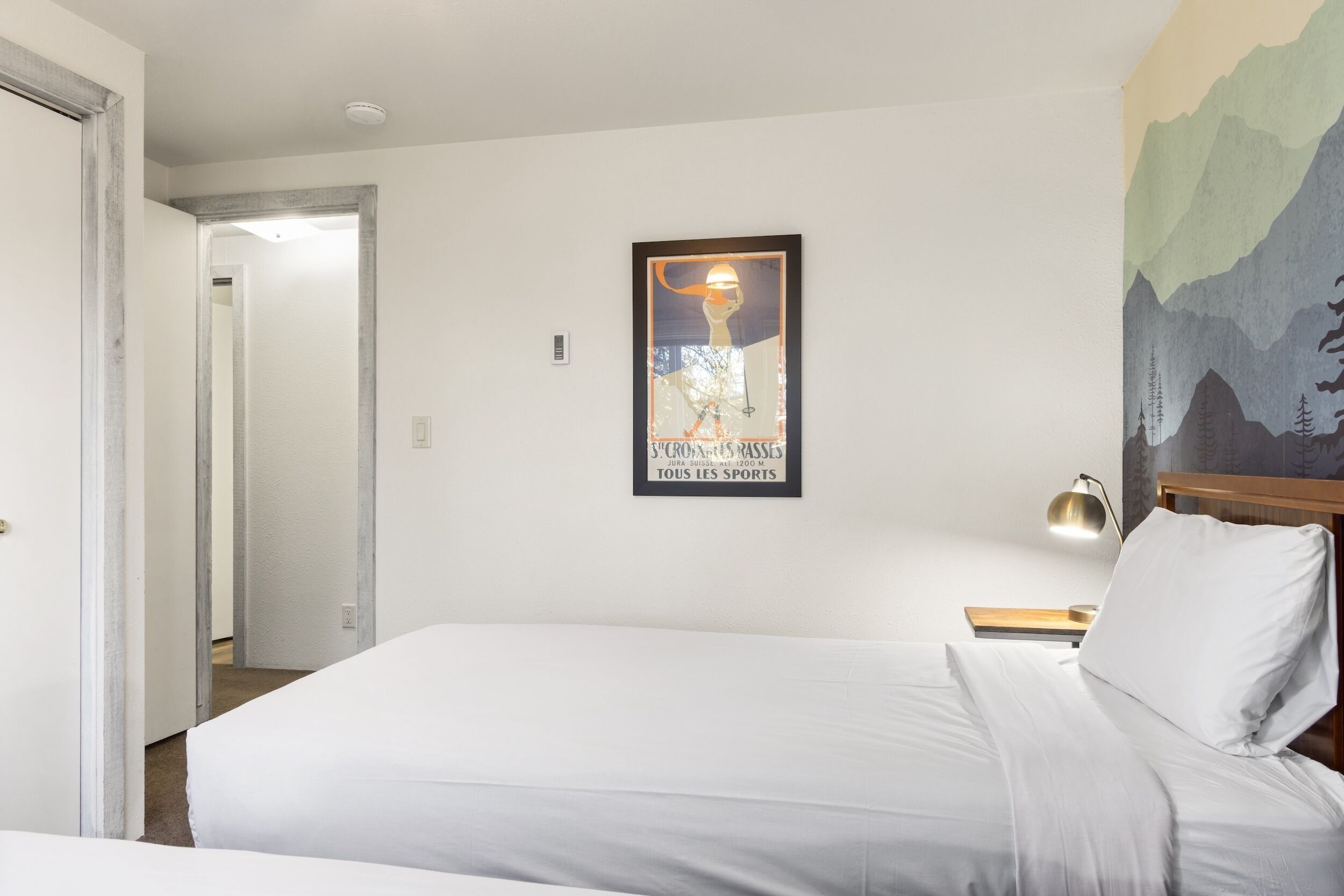 Ein Hotelzimmer mit einem Bett, einem Nachttisch mit Lampe, einem gerahmten Poster an der Wand und einer Tür, die in einen anderen Raum führt.