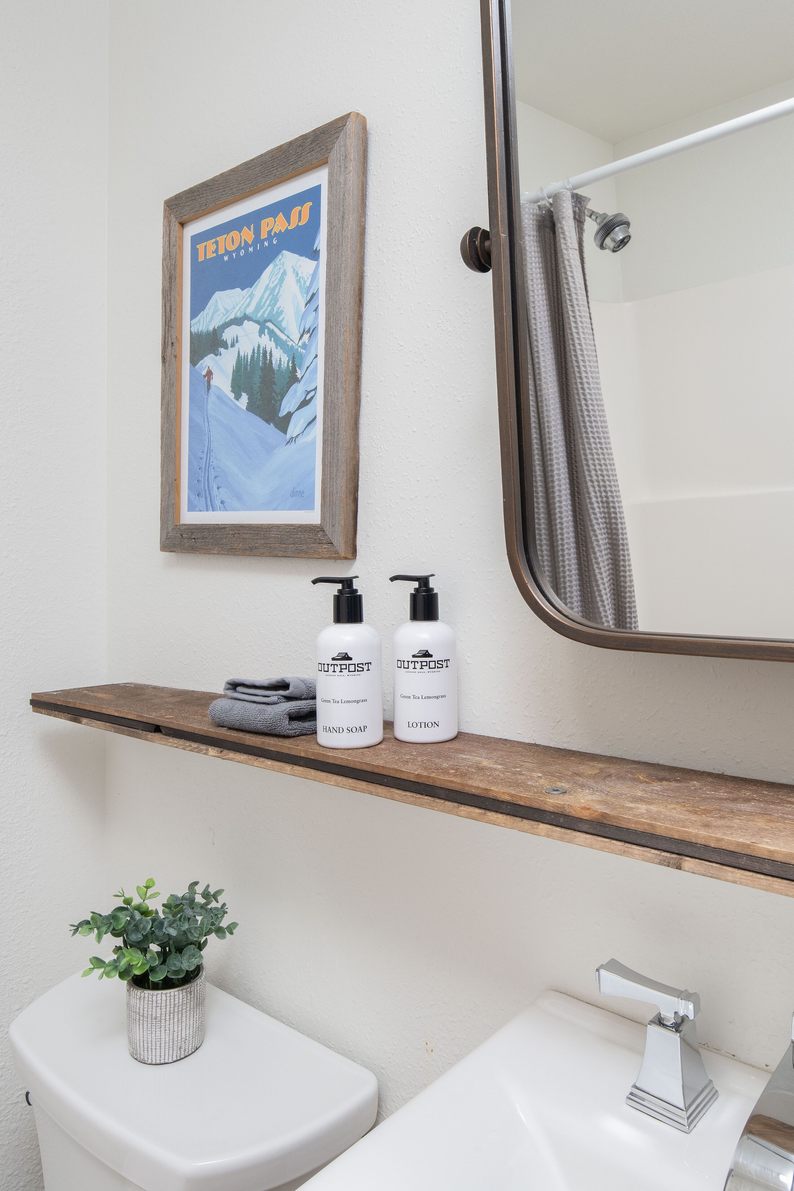 Een badkamer met een houten plank waarop toiletartikelen staan, een lijst met een berglandschap, een potplant en een witte wastafel met een moderne kraan.