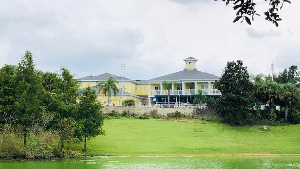 A large building with a yellow facade and a grey roof, surrounded by lush greenery and a body of water.