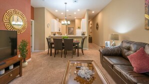 A living room with a brown leather sofa, a coffee table with decorative items, a TV stand, and a dining area with chairs and a kitchen in the background.