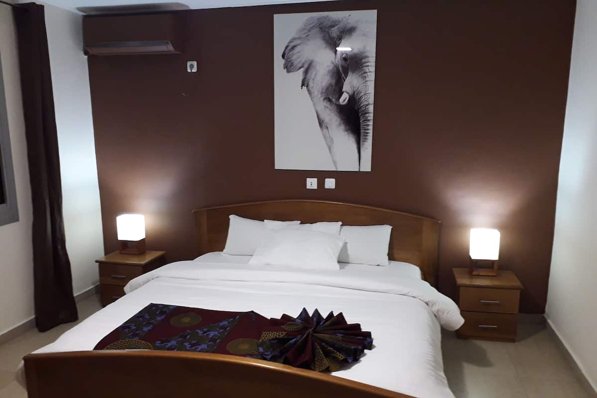 A neatly made bed with white linens and a decorative pillow, flanked by two bedside tables with lamps, in a room with brown walls and a large elephant artwork above the bed.