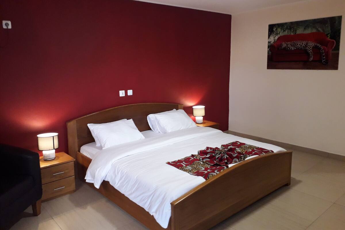 A hotel room with a large bed, two bedside lamps, a nightstand, and a red wall.