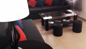 Een moderne woonkamer met een zwarte bank, rode en blauwe kussens, een glazen salontafel en zwarte poefs.