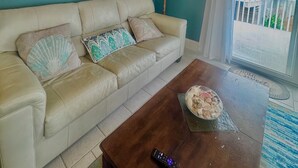 Una sala de estar con paredes verde azulado, un sofá color crema, una mesa de centro de madera y un cuadro con una escena de playa.