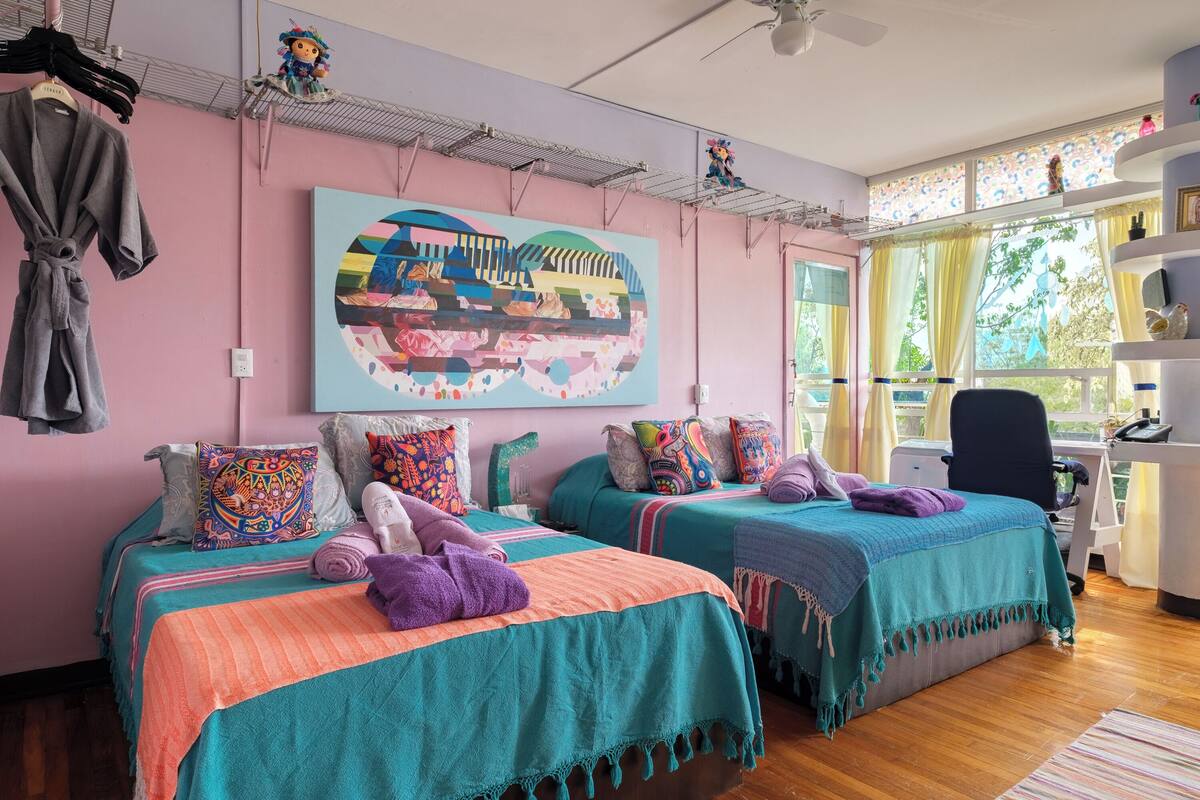 A bedroom with two beds, colorful bedding, a large abstract painting, and a desk with a chair.