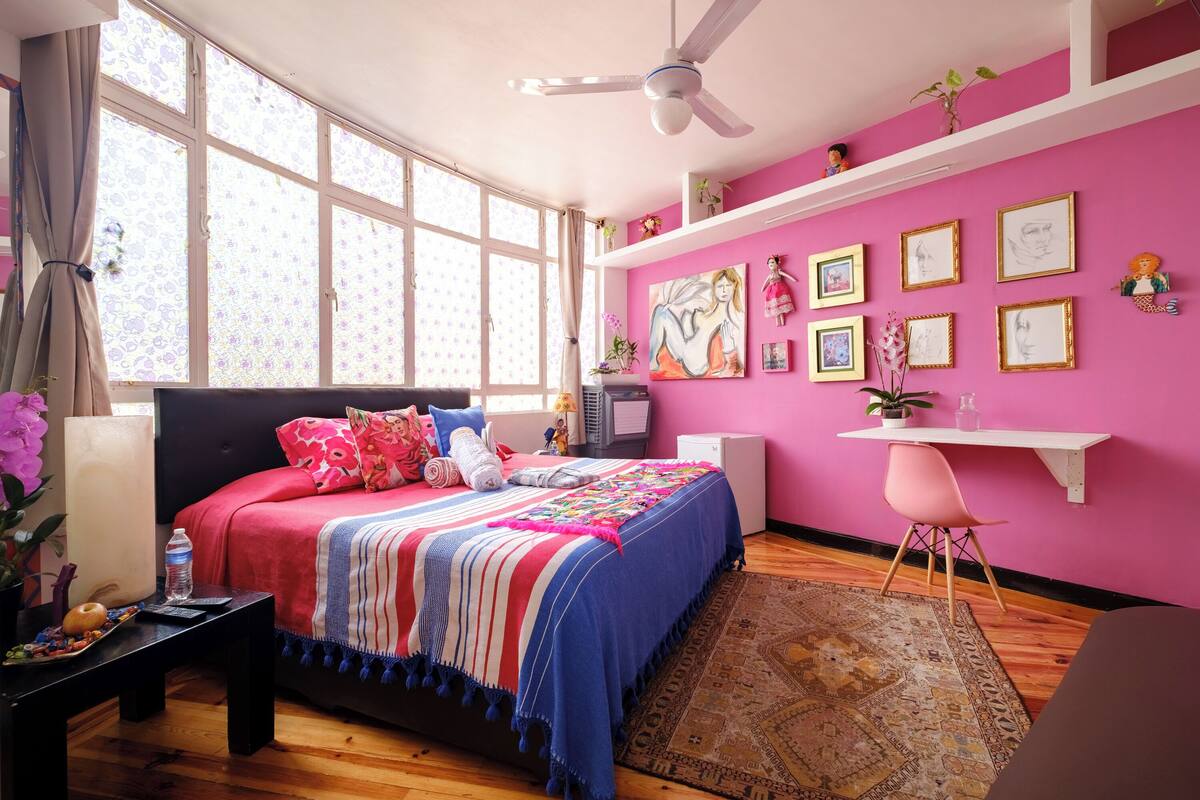 A bedroom with a large bed, a desk, a chair, a ceiling fan, and a window with curtains.