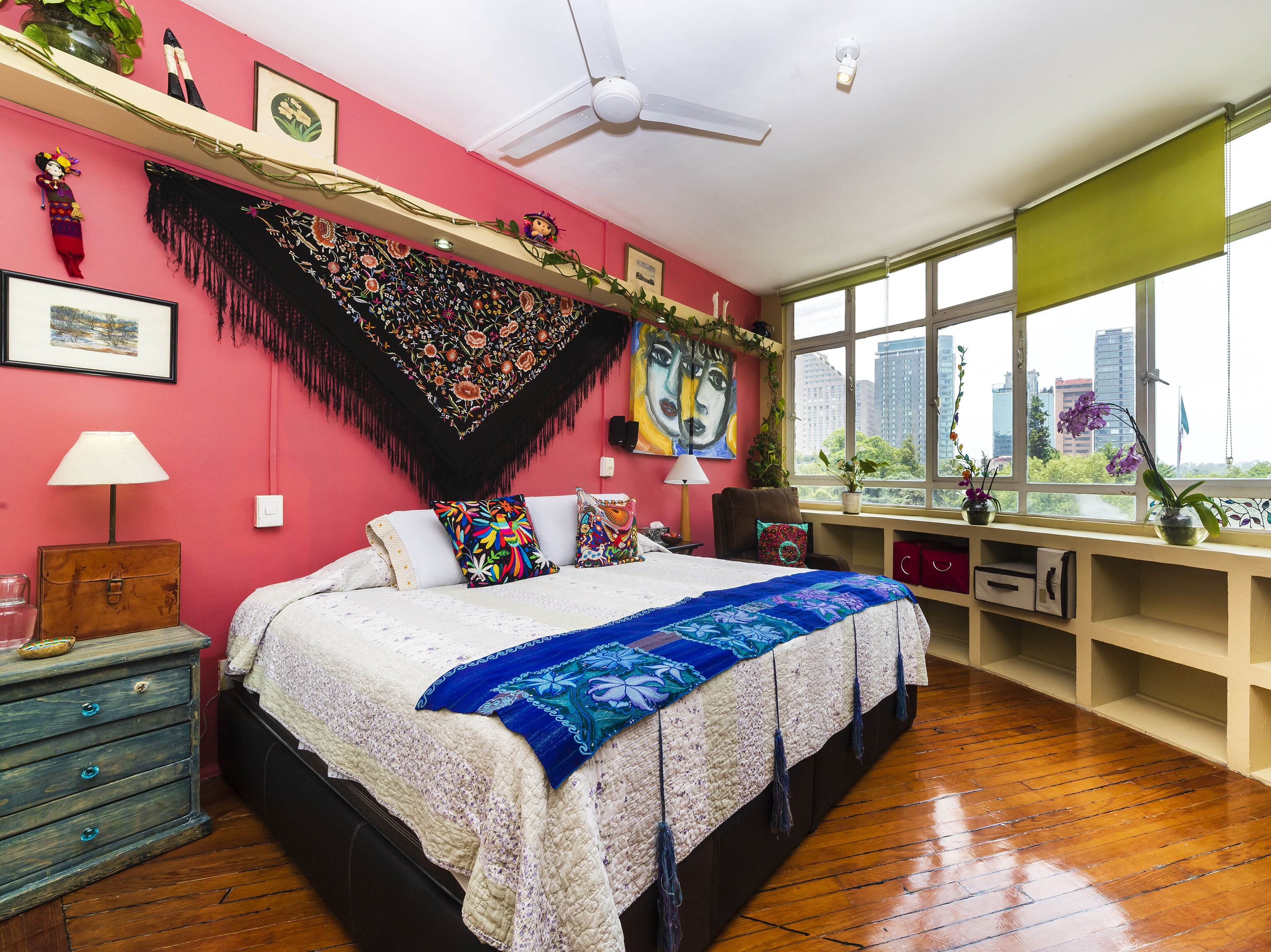 A bedroom with a large bed, a desk with a computer, a window with a city view, and a ceiling fan.