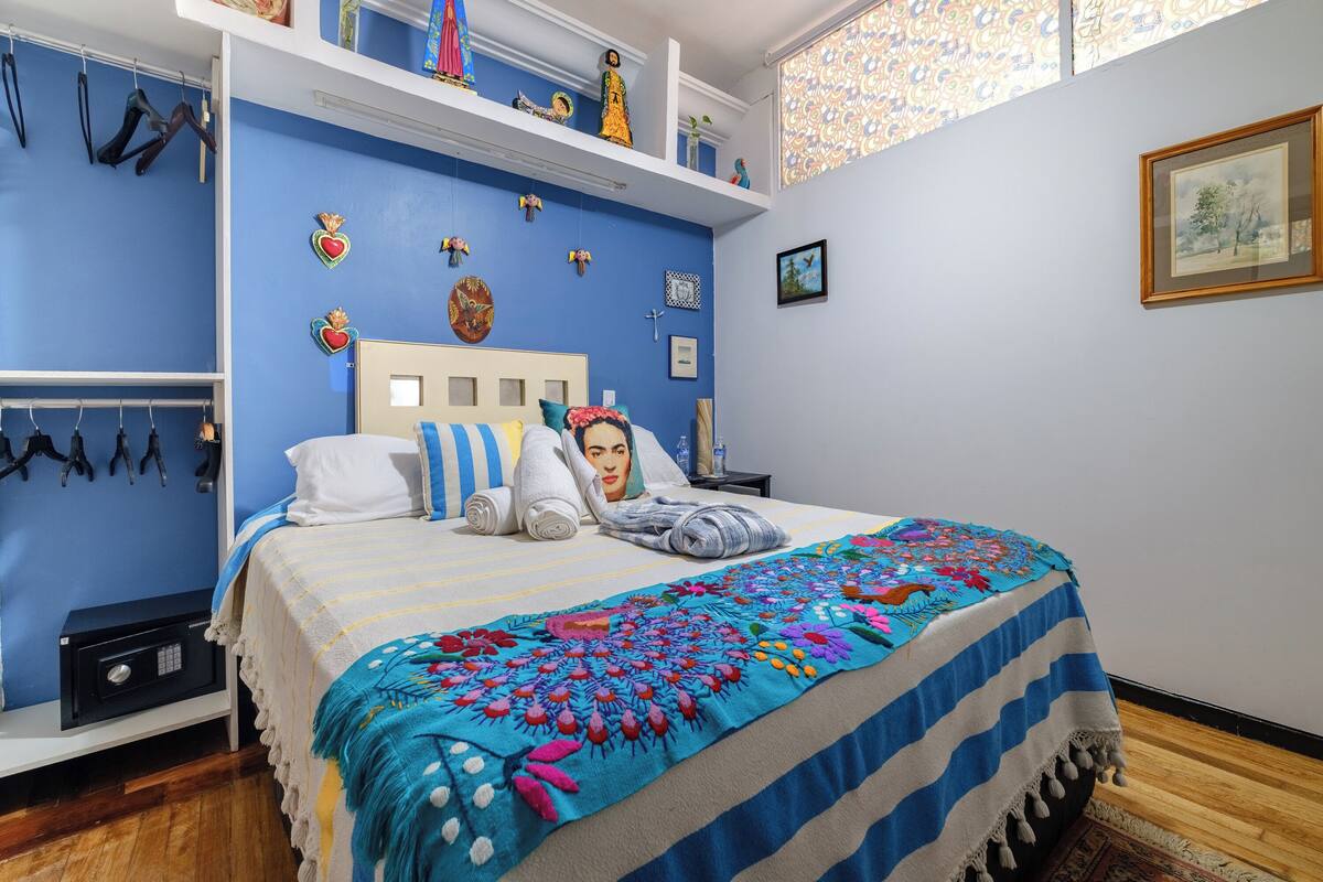 A bedroom with a bed, a nightstand, a wall-mounted shelf with decorative items, and a window with patterned curtains.