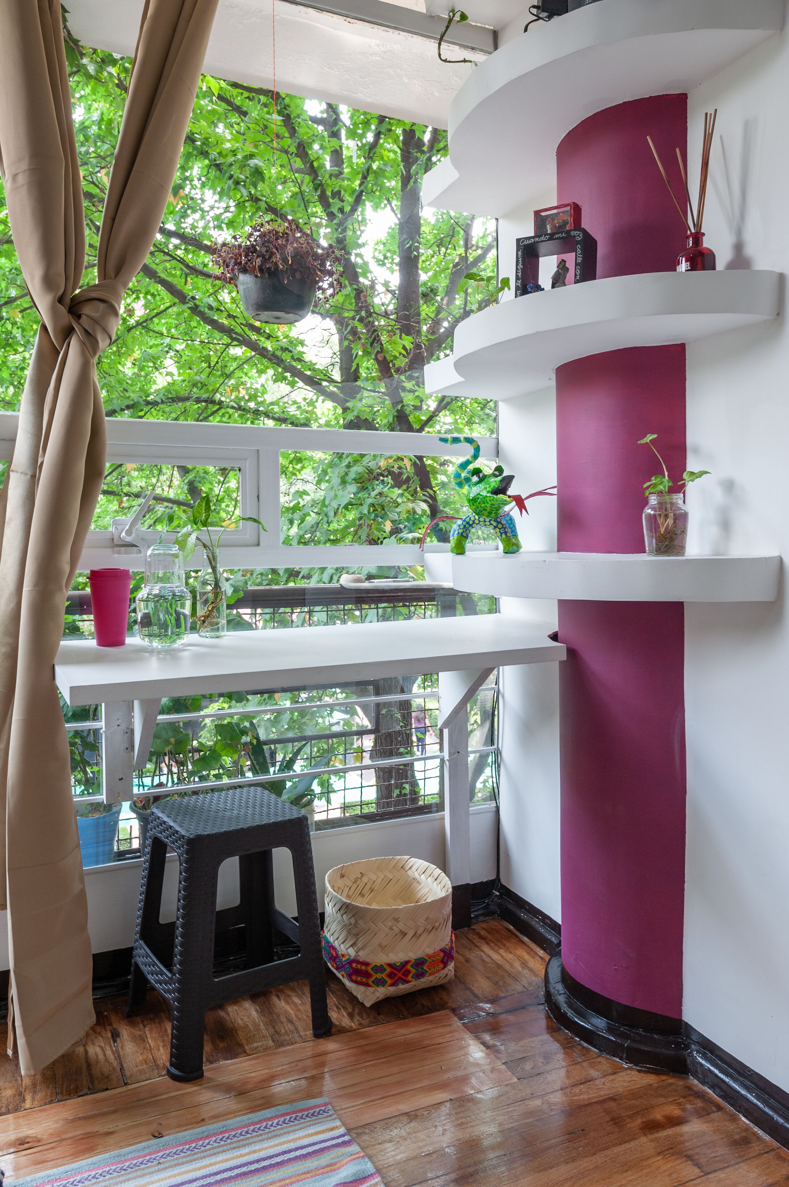 A room with a wooden floor, a black stool, a woven basket, a pink cup, a hanging plant, and a shelf with a vase and a picture frame.