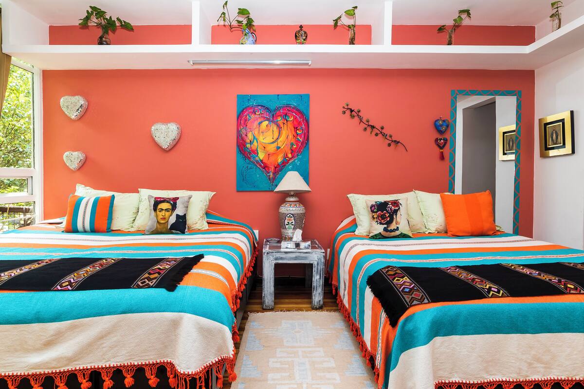A room with two beds, a heart-shaped wall decoration, a colorful painting, and a small table with a lamp.