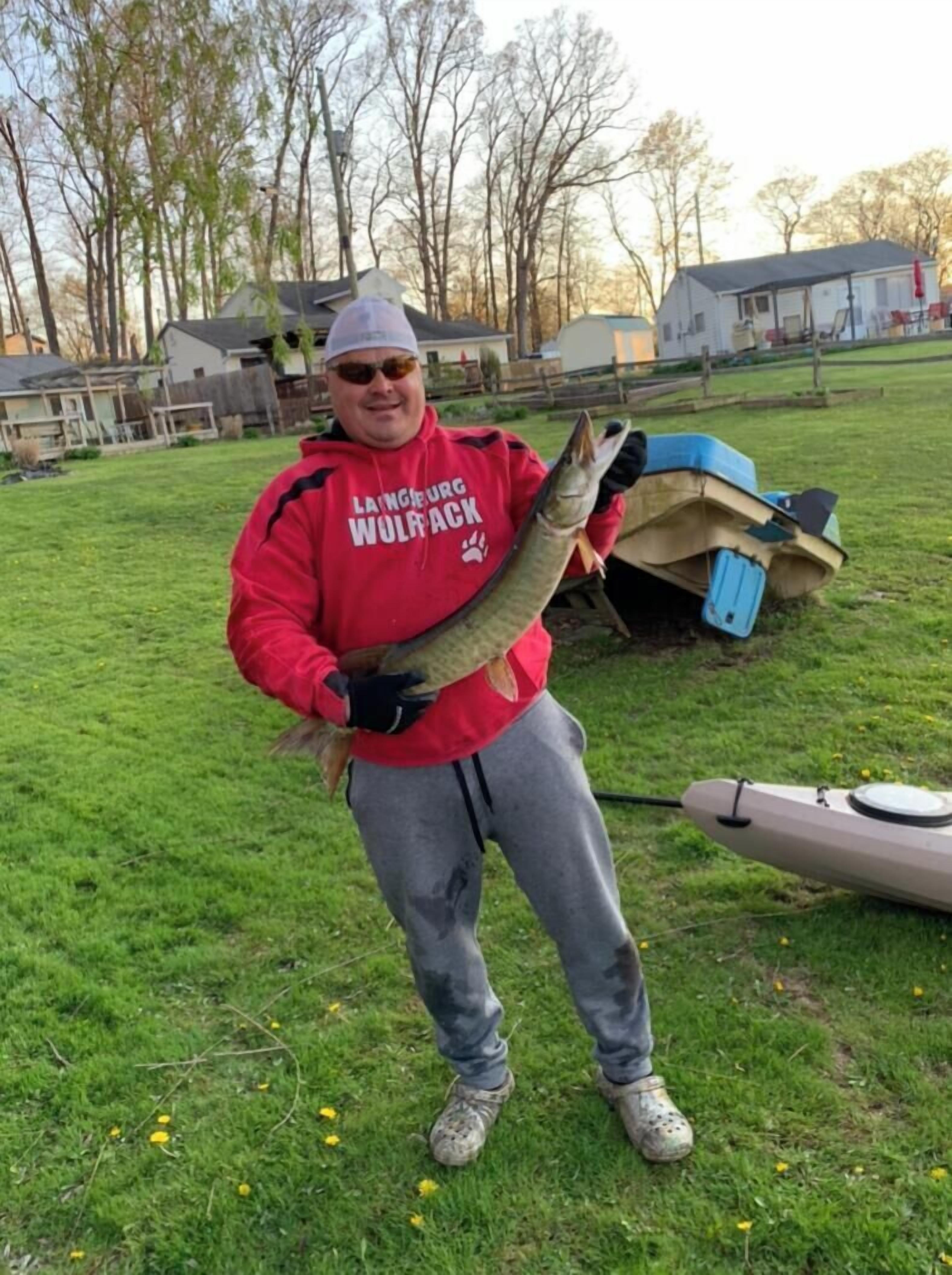 A person holding a fish in a grassy area with a kayak and a house in the background.