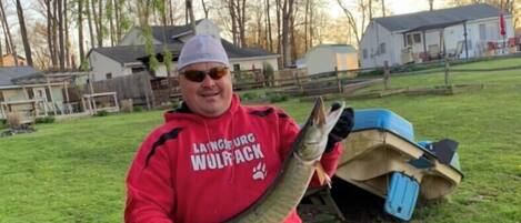 A person holding a fish in a grassy area with a kayak and a house in the background.