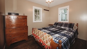 Un dormitorio con un aparador de madera, una cama con colcha escocesa, un ventilador de techo y dos ventanas.