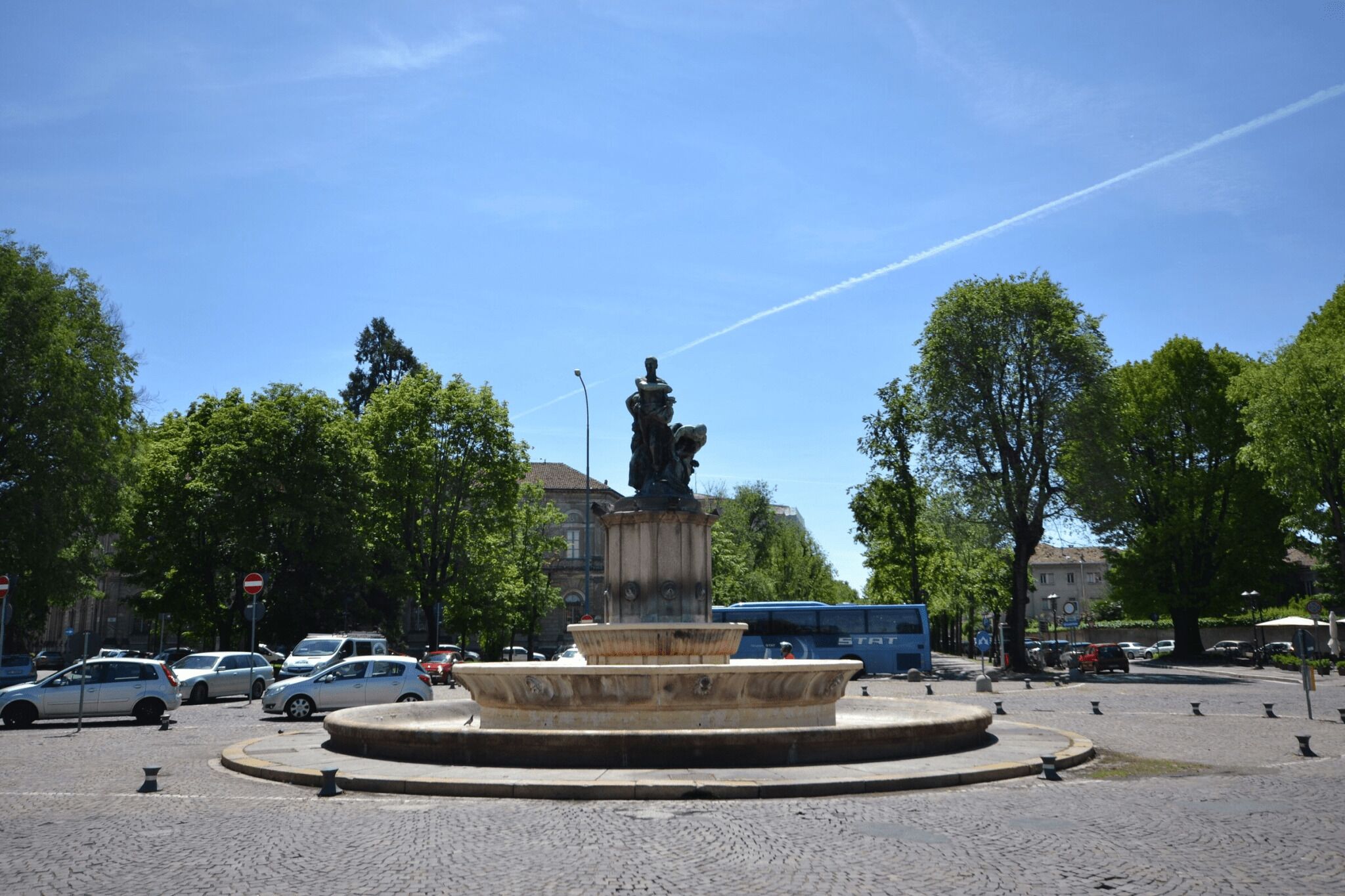 Una glorieta con una fuente en el centro, rodeada de árboles y coches aparcados.