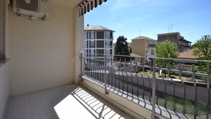 Ein Balkon mit Metallgeländer, einer weißen Klimaanlage und Blick auf Gebäude und B äume im Hintergrund.