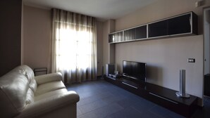 Ein Wohnzimmer mit einer Couch, einem Flachbildfernseher, einem Regal über dem Fernseher und einem Fenster mit Vorhängen.